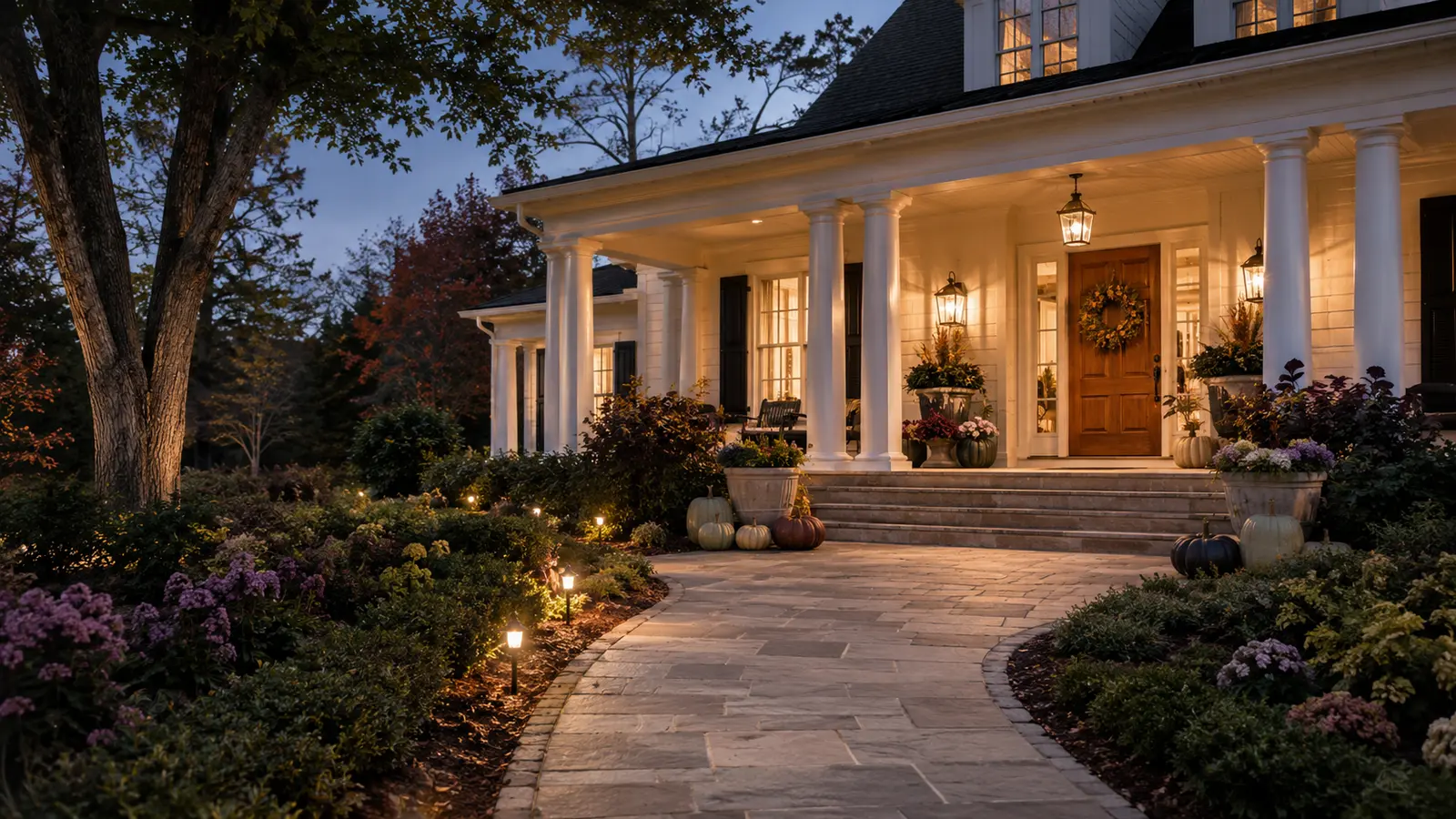 A guest-eye view from the driveway following a broad illuminated walkway toward an unobstructed, warmly styled fall porch